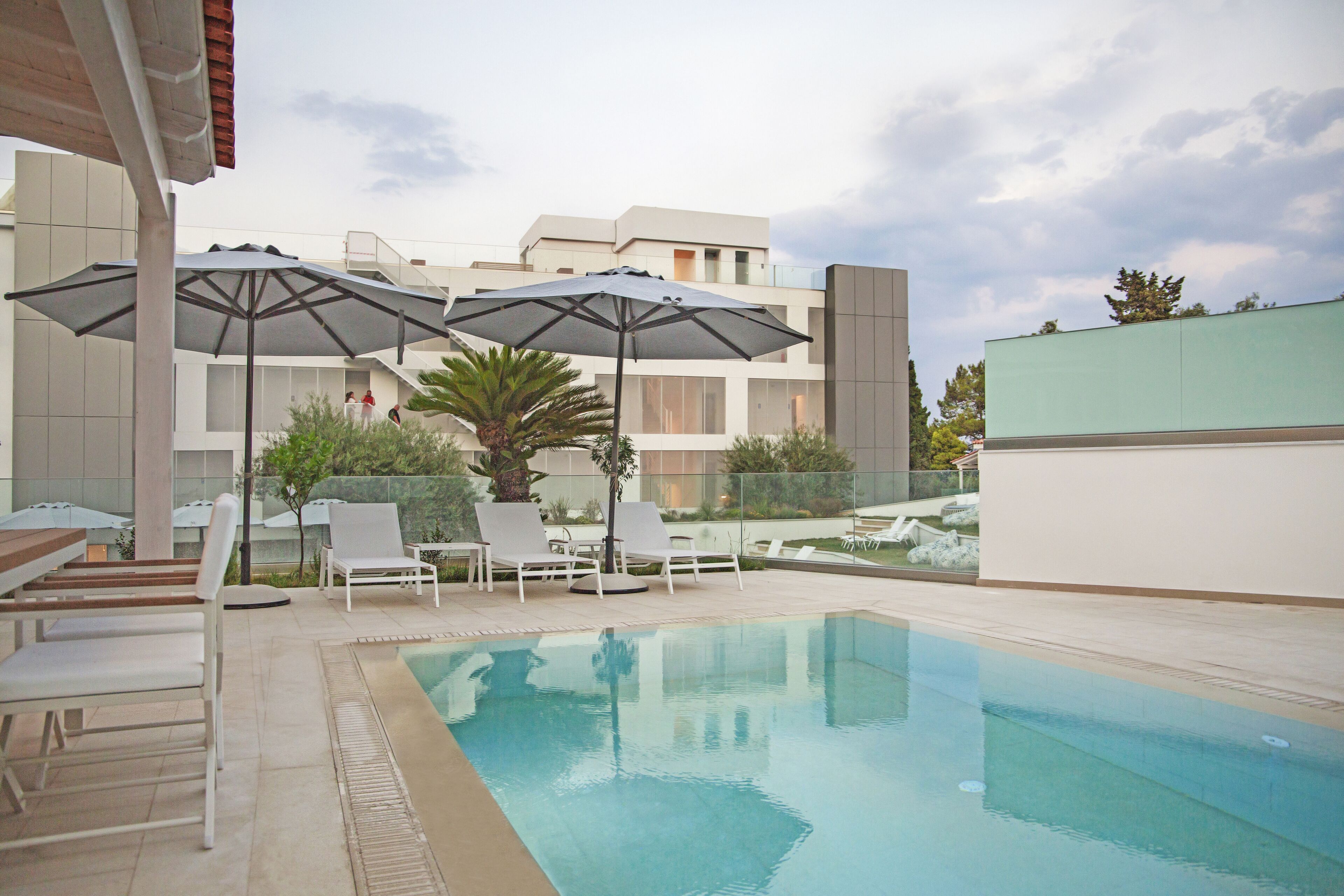 Zona de piscina con sillones de descanso y sombrillas, rodeada por un edificio moderno con balcones.