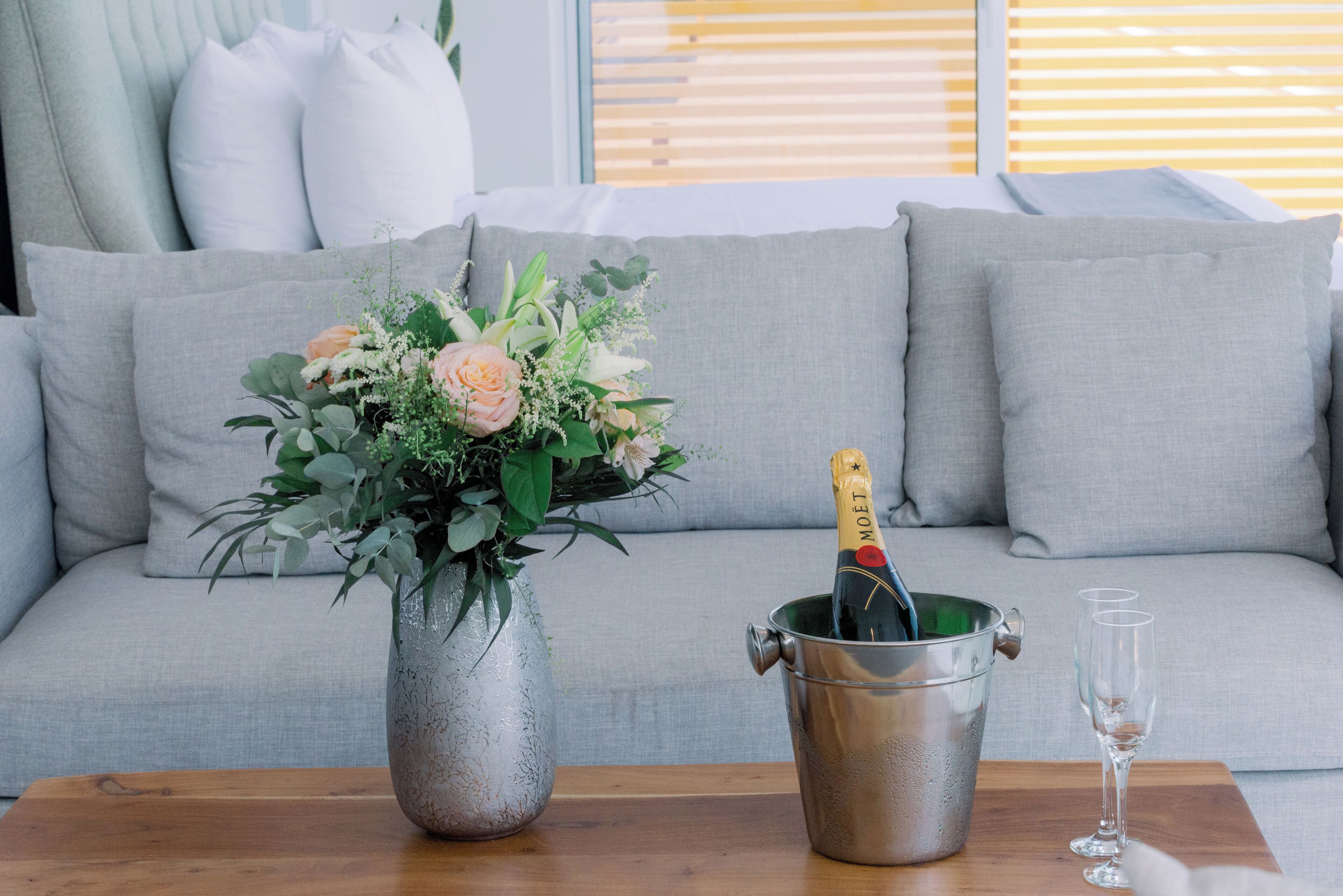 A living room with a grey sofa, a metal bucket with a bottle of champagne, and a vase with flowers on a wooden table.