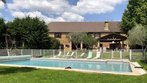 Ein Schwimmbecken mit Liegestühlen, ein Steinhaus mit einem Ziegeldach und ein eingezäunter Garten.