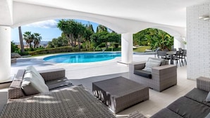 A modern outdoor lounge area with wicker furniture, a pool, and a view of palm trees and a cityscape. - Villa Catalina (Benalmádena)