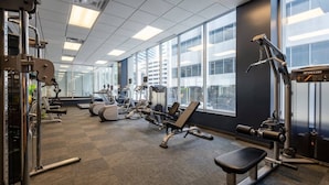 A gym with various exercise machines, large windows, and a view of buildings outside. - CozySuites - Remi (Dallas)