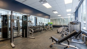 A gym with various exercise equipment, large windows, and a mirror wall. - CozySuites - Remi (Dallas)