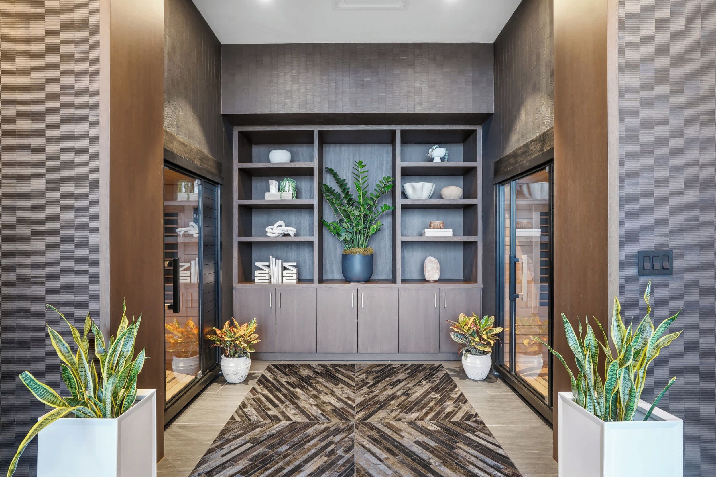 A modern hallway with a geometric patterned rug, grey cabinets, and potted plants.