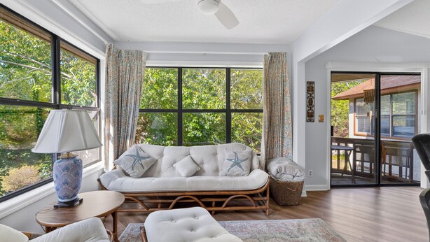 A room with a sofa, ottoman, and a table with a lamp. There are large windows with curtains, a ceiling fan, and a view of trees outside. - 2br/2ba Walk to pier/village area.  1/2 mile to beach (Saint Simons Island)