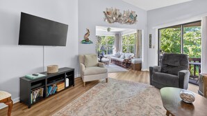 A living room with a flat-screen TV, a sofa, a chair, a bookshelf, and a large window. - 2br/2ba Walk to pier/village area.  1/2 mile to beach (Saint Simons Island)