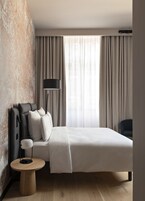 A modern hotel room with a large bed, a bedside table, a lamp, and a view of the outdoors through sheer curtains.
