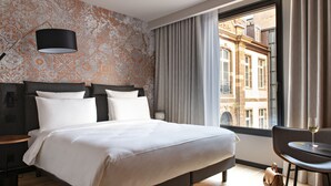 Una camera d'albergo con un letto grande, un comodino, una lampada, una finestra con tende e vista sugli edifici.