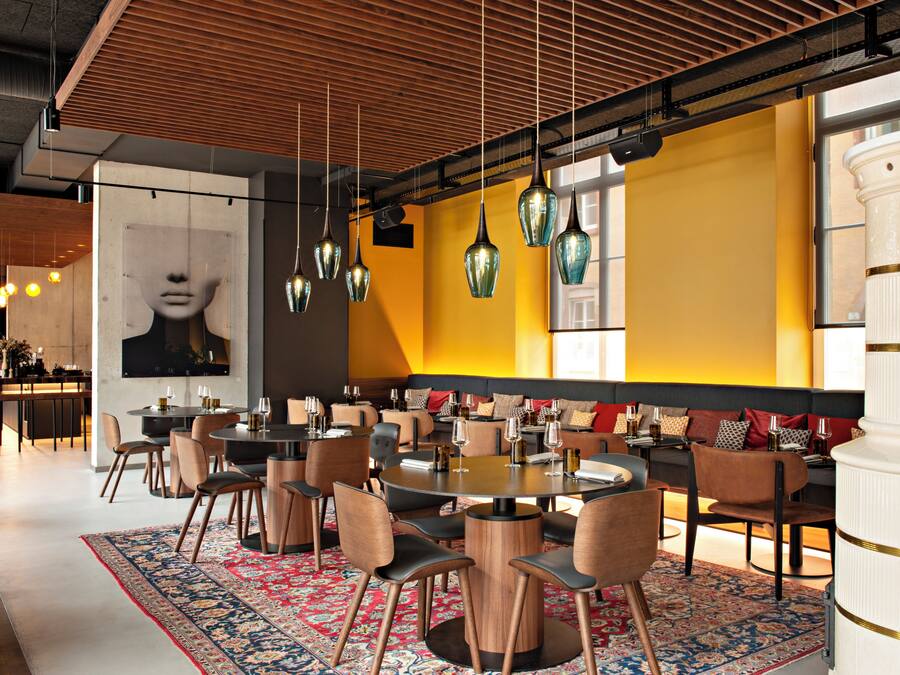 Un restaurante moderno con mesas redondas, sillas, lámparas colgantes y un gran tapiz decorativo.