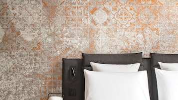 Un dormitorio con papel tapiz estampado, una cama con ropa de cama blanca, una mesita de noche con lámpara y un tocador.