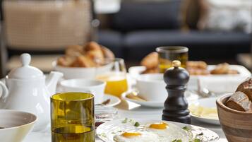 Une table de petit-déjeuner avec divers plats, notamment des fruits, des œufs et des viennoiseries, dans une pièce bien éclairée, avec un canapé et une fenêtre en arrière-plan.