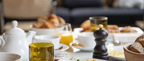 Une table de petit-déjeuner avec divers plats, notamment des fruits, des œufs et des viennoiseries, dans une pièce bien éclairée, avec un canapé et une fenêtre en arrière-plan.