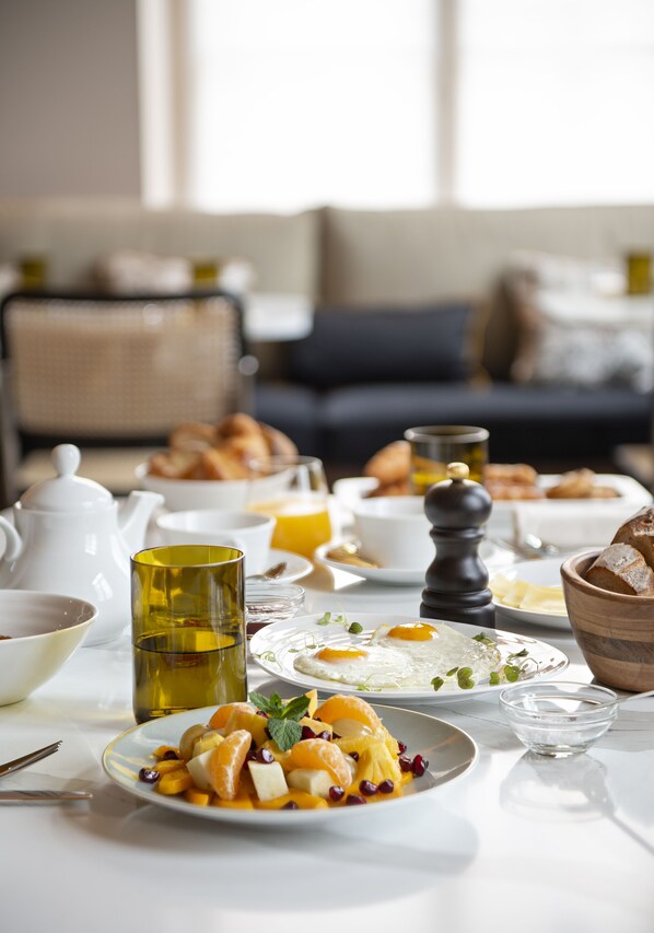 Una mesa de desayuno con diversos platos, como frutas, huevos y pasteles, en una habitación luminosa con un sofá y una ventana al fondo.