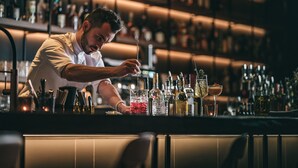 Un barista prepara cocktail dietro un bancone con scaffali pieni di bottiglie sullo sfondo.
