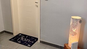 A hotel room with a white door, a doormat with the word "Main" on it, a wall-mounted light fixture, and a painting of a beach scene. - Maritime vacation apartment in Wilhelmshaven on the North Sea (Wilhelmshaven)