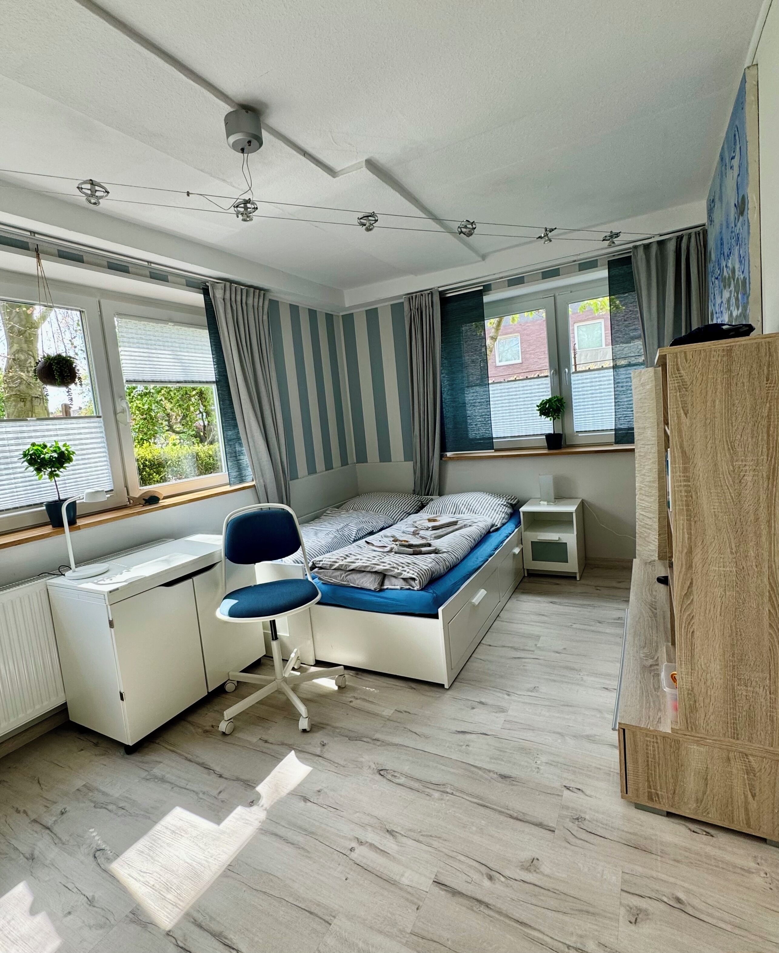 Une chambre à coucher comprenant un lit, un bureau, une chaise, un radiateur, des fenêtres, des rideaux et une armoire en bois.