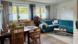 A modern living room with a blue sofa, wooden dining table, and chairs. There are large windows with curtains, a potted plant, and a small table with a remote control. - Maritime vacation apartment in Wilhelmshaven on the North Sea (Wilhelmshaven)