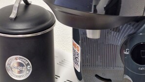 Une machine à café, une cafetière noire et deux dosettes Tassimo sur une surface blanche.