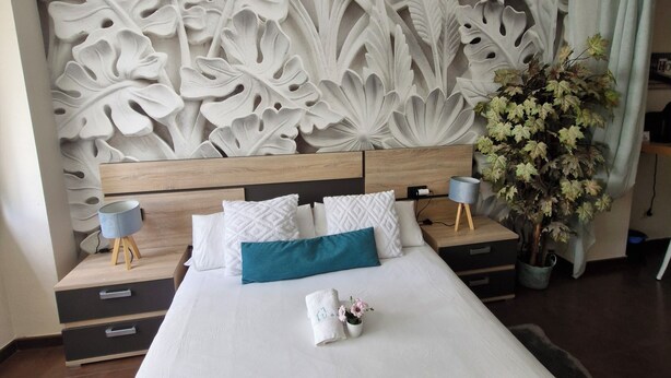 Une chambre d’hôtel moderne avec un grand lit, un panneau décoratif au mur, une table de chevet, une lampe, une plante en pot et une petite table sur laquelle est posé un vase.