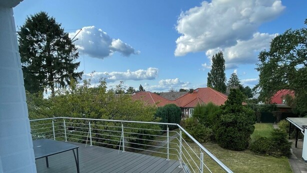 Una terraza de madera con barandillas de metal, con vista a una zona residencial con casas y árboles bajo un cielo parcialmente nublado.
