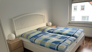 Una cama blanca con cabecera, dos mesitas de noche y una ventana con vistas a edificios.