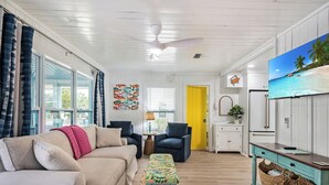 A spacious living room with a beige sofa, a blue armchair, and a colorful ottoman. There is a television mounted on the wall, a ceiling fan, and a yellow door leading to another room. - Sassy Seagull -Classic beach cottage, moments from the sand, beautiful pool! (Anna Maria)