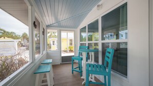 A covered porch with teal chairs and white benches, large windows, and a view of the outside. - Sassy Seagull -Classic beach cottage, moments from the sand, beautiful pool! (Anna Maria)