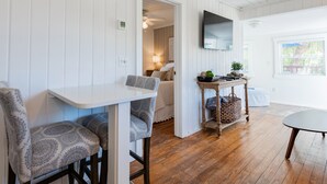 Una habitación con pared de madera blanca, ventilador de techo, televisor montado en la pared, mesa de madera con una cesta y ventana con persianas.