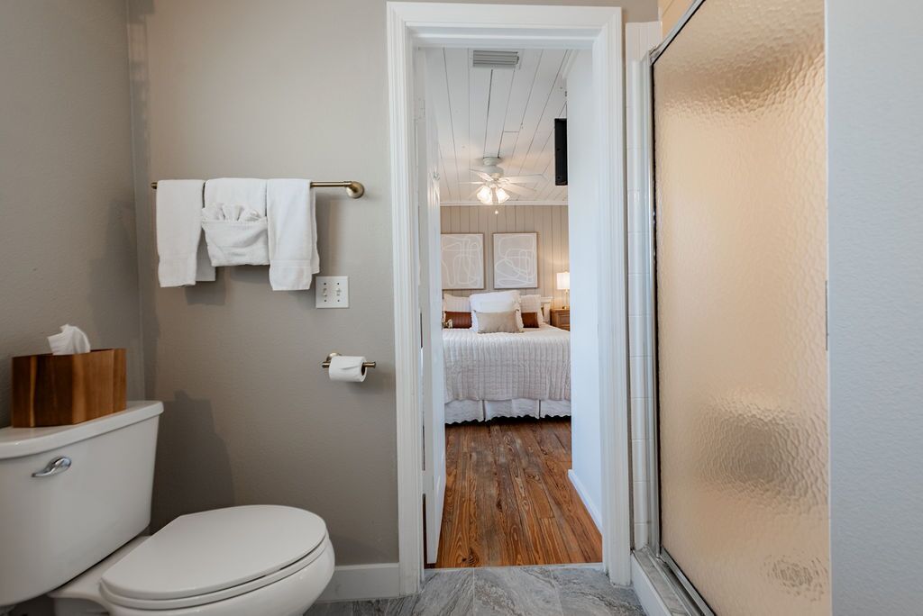 A bathroom with a toilet, towel rack, and a wooden floor leading to a bedroom with a bed and a ceiling fan.
