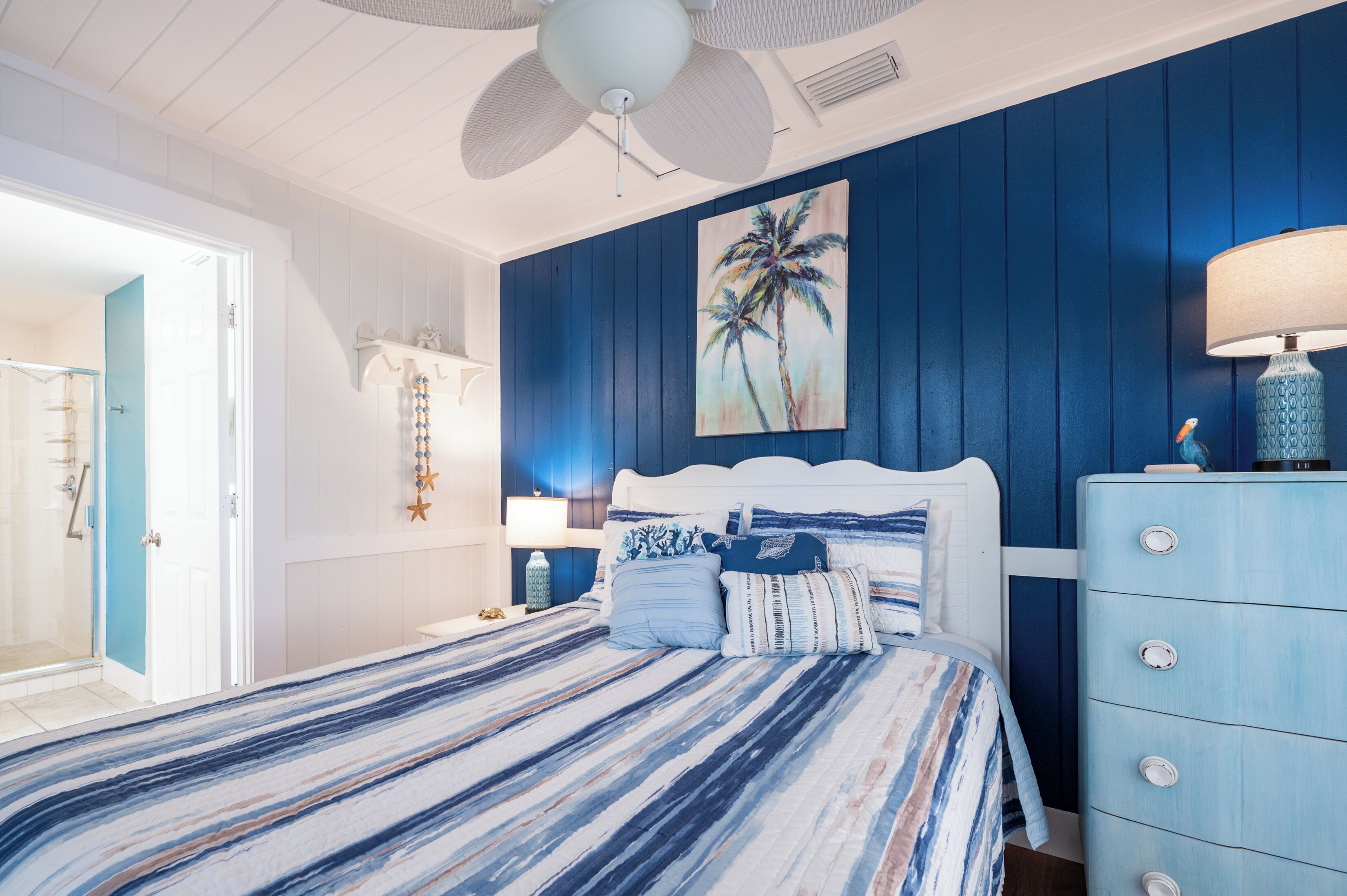 A bedroom with a bed, a nightstand, a dresser, a ceiling fan, and a painting of palm trees.