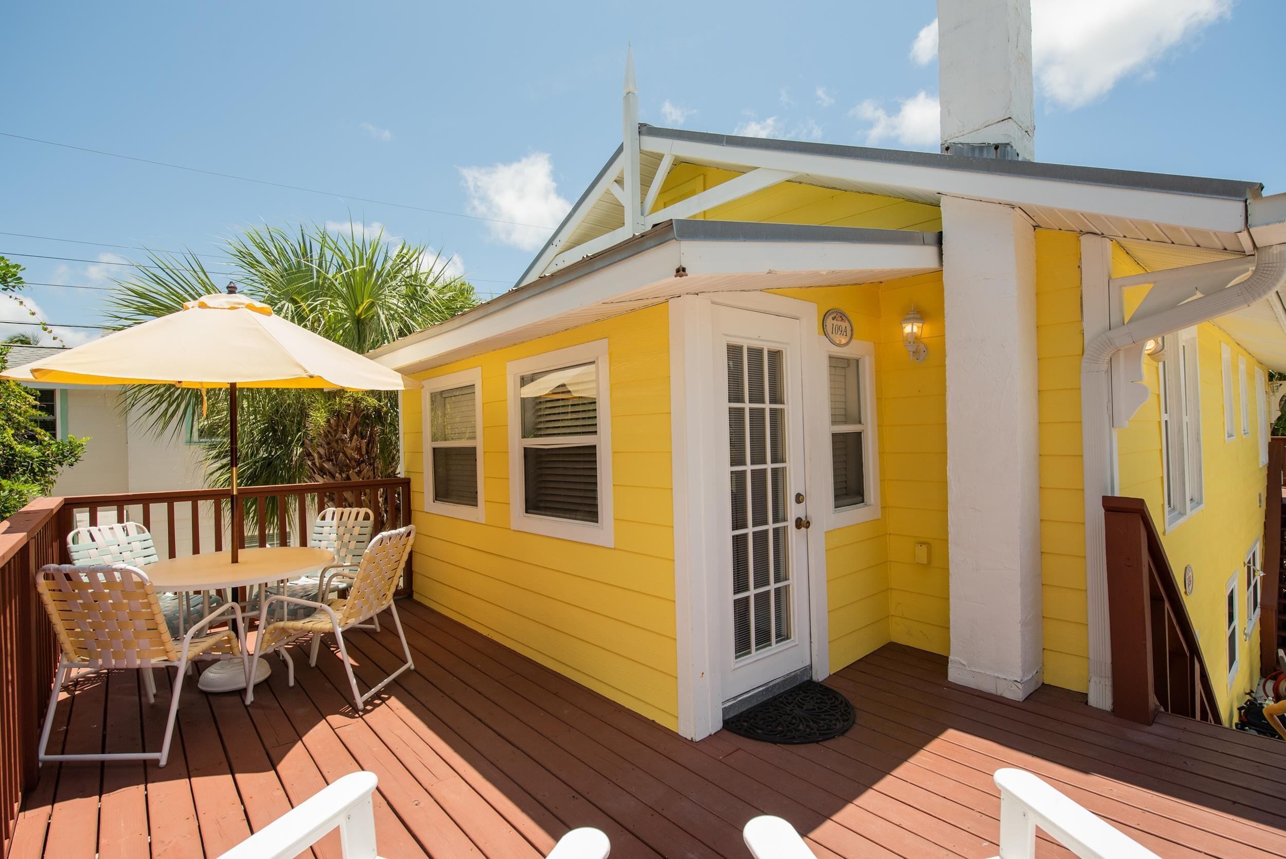Une maison jaune avec une terrasse en bois, une table à manger avec des chaises et un patio couvert.