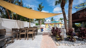An outdoor patio with wooden furniture, a yellow shade sail, palm trees, and a view of a building with colorful umbrellas.