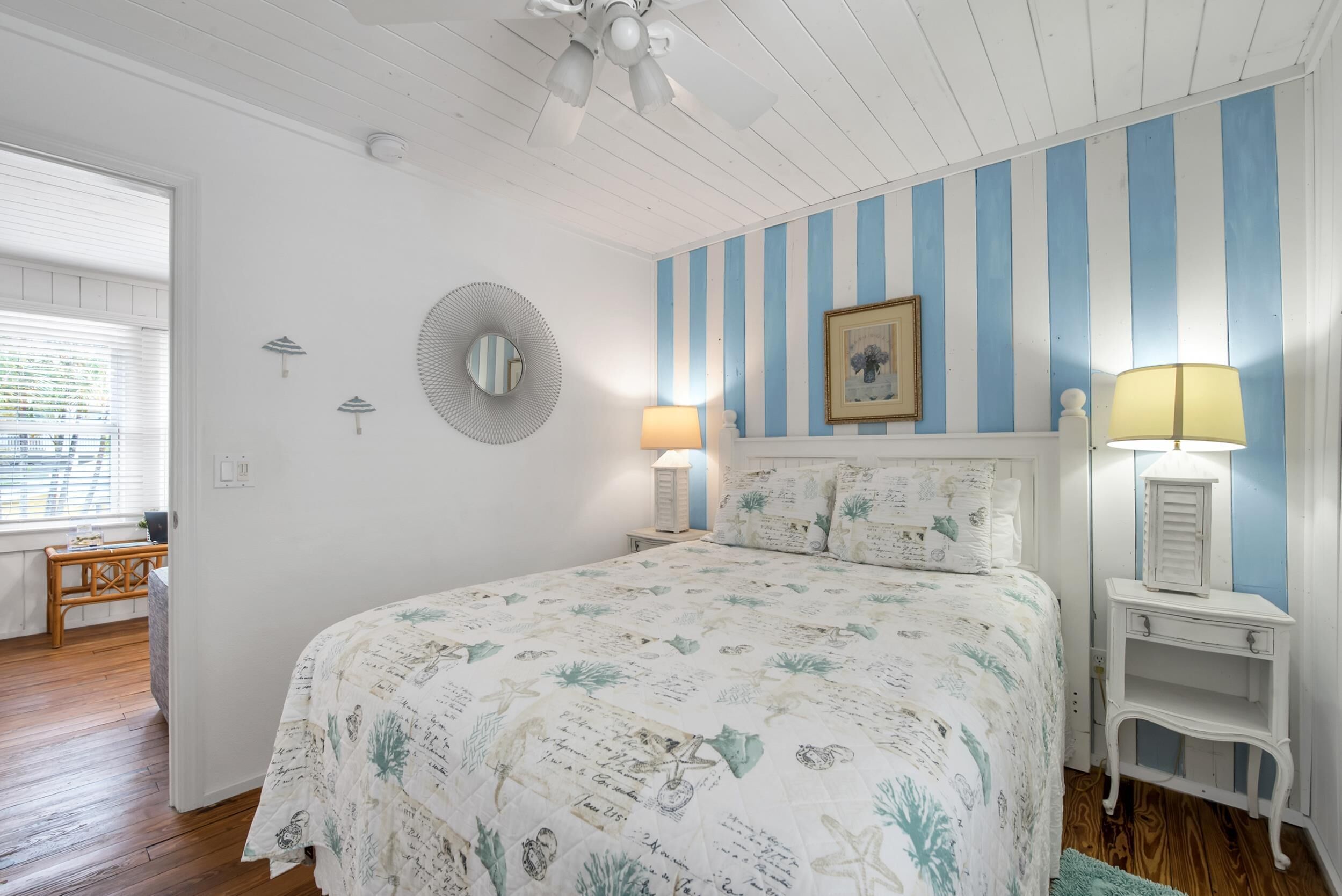A bedroom with a bed, two bedside lamps, a wall clock, a mirror, and a window with blinds.