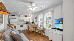 A spacious living area with a gray sofa, wooden dining table, and chairs, a kitchenette, and a TV displaying a beach scene.