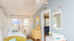 A bedroom with a bed, a yellow dresser, a white nightstand, a mirror, and a window with blinds. - Anna Maria Beach Cottage 110  Traditional beach cottage, moments from the sand! (Anna Maria)