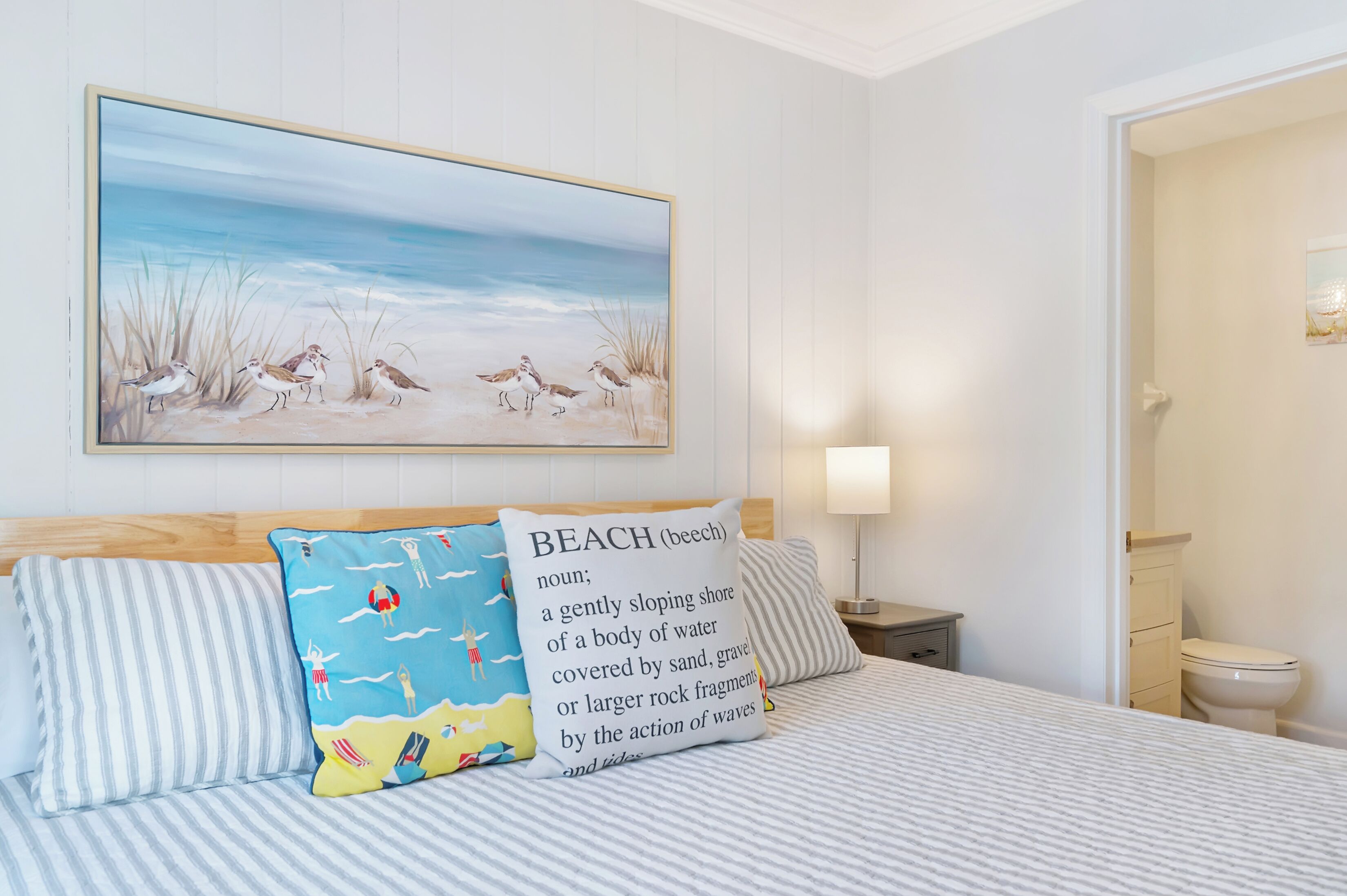 Une chambre à coucher avec un lit, des oreillers, un tableau représentant des oiseaux accroché au mur et une table de chevet avec une lampe.