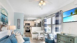 Un salón luminoso con un sofá azul, una mesa de comedor blanca y una cocina al fondo.