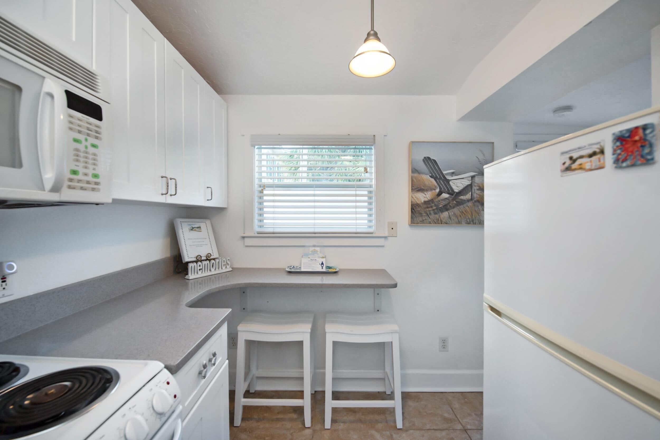 A kitchen with white cabinets, a microwave, a stove, two white bar stools, a window with blinds, and a hanging light fixture.