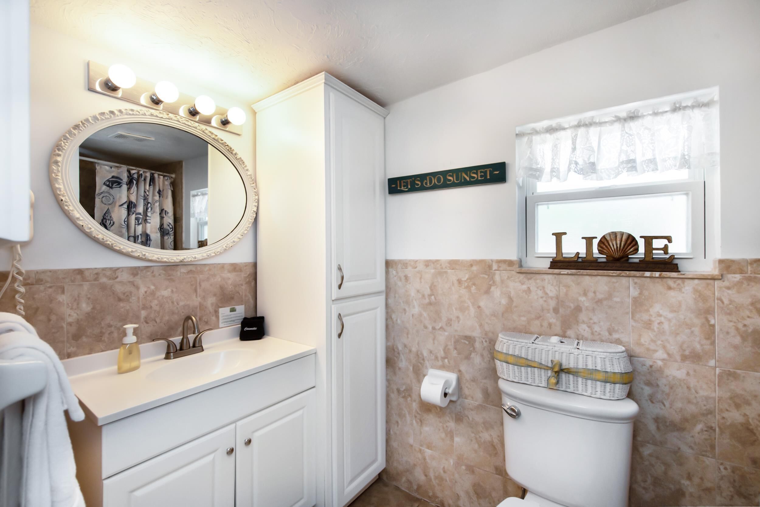 A bathroom with a vanity, a mirror with lights, a toilet, and a sign that reads "LET'S DO SUNSET".