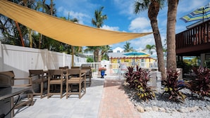 An outdoor patio with wooden furniture, a yellow shade sail, palm trees, and a view of a building with colorful umbrellas. - Anna Maria Beach Cottage 110G  Updated beach cottage, with pool and beach gear! (Anna Maria)