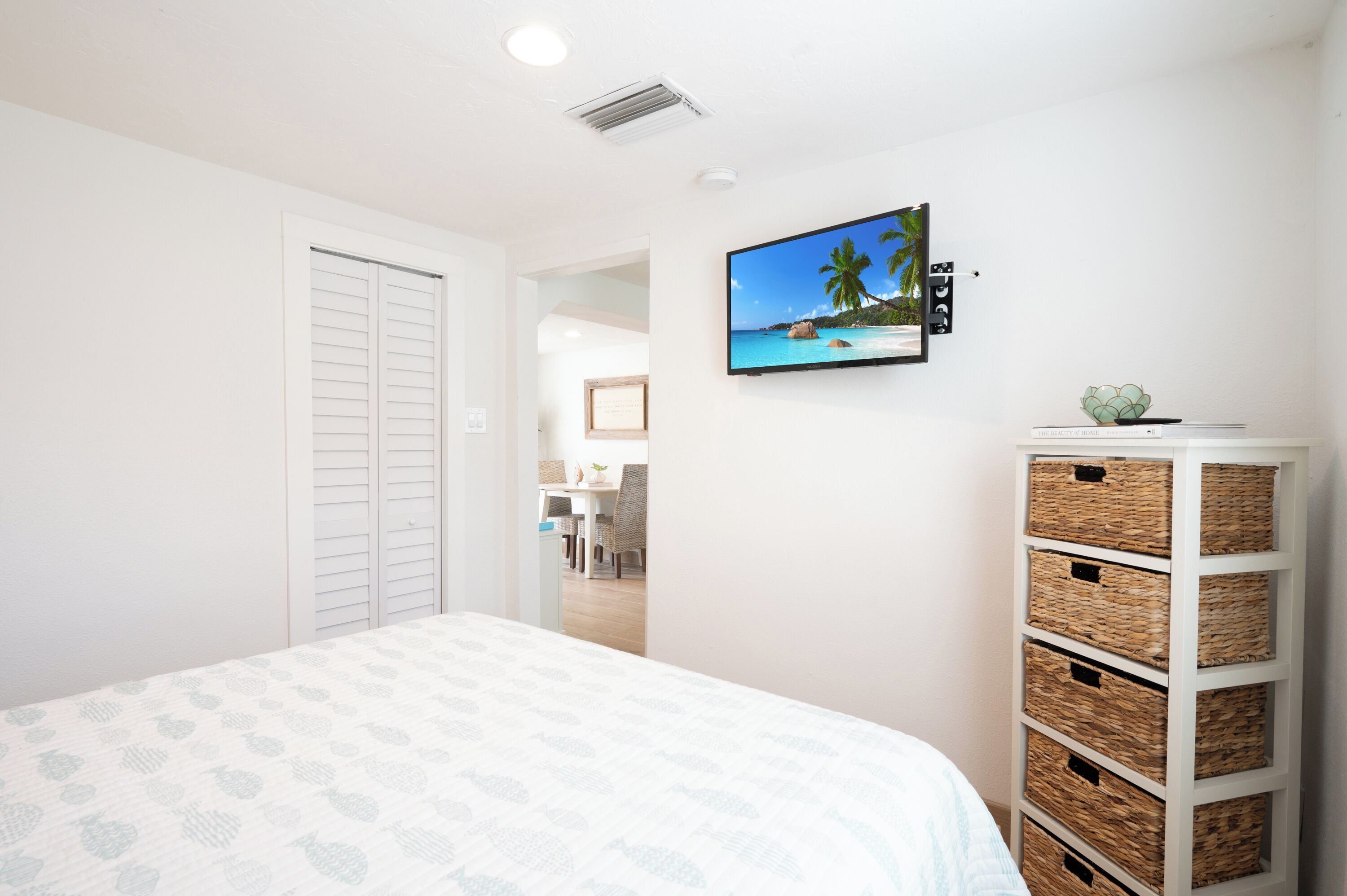 A bedroom with a bed, a TV on the wall, a white wardrobe, and a wicker storage unit.