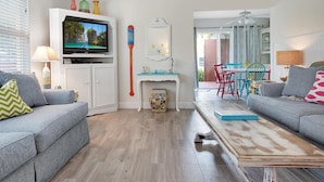 A living room with a television, a fireplace, a dining area with colorful chairs, and a balcony with a view. - Flirty Flamingo Cottage Updated beach cottage, moments from the sand, w/pool! (Anna Maria)