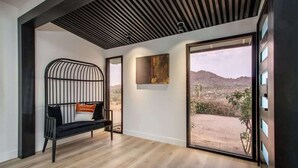 A modern interior with a wooden floor, a black metal bench with cushions, a large window with a view of a desert landscape, and a painting on the wall. - @ Marbella Lane - The Wine Mine Desert Escape (Joshua Tree)