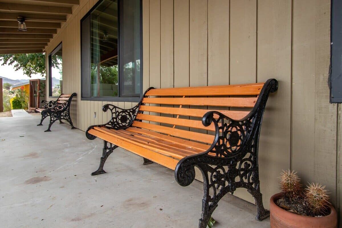 A wooden bench with ornate black metal legs on a concrete floor, a potted cactus, and a building with vertical siding and large windows.