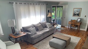 Un salon avec un canapé gris, une table basse en bois, un fauteuil gris, une lampe et une porte donnant sur l’extérieur.