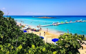 A beach with clear turquoise water, people swimming and sunbathing, and boats docked near the shore. - 5 Star Apartment for Rent in Cyprus, Protaras Apartment 1210 (Paralimni)
