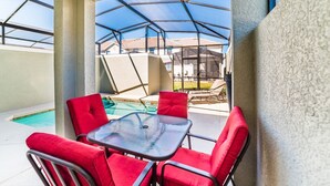 Eine überdachte Terrasse mit einem Glastisch und roten Stühlen, mit Blick auf einen Swimmingpool und einen Garten mit Gartenmöbeln.