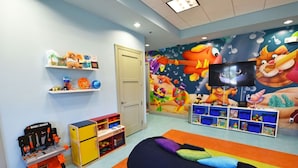 Une salle de jeux pour enfants avec une fresque murale colorée, des étagères remplies de jouets, un fauteuil en forme de sac et un téléviseur.