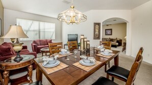 Un comedor con una mesa de madera dispuesta para cuatro personas, rodeada de sillas. Hay una araña sobre la mesa y un televisor montado en la pared.