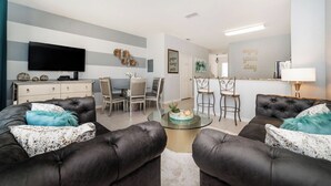 A modern living room with a flat-screen TV, two leather sofas with decorative pillows, a glass coffee table, and a dining area with a chandelier. - 5 Star Townhome on Windsor Hills Resort With Large Private Pool, Orlando Townhome 4922 (Kissimmee)