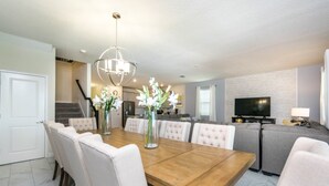 A modern dining area with a wooden table and white upholstered chairs, a chandelier, and a kitchen in the background. - Luxury Mansion on Storey Lake Resort, Minutes From Disney World, Orlando Mansion 2672 (Kissimmee)